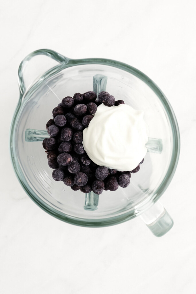 frozen blueberries and plain greek yogurt in a glass blender on a white marble countertop.