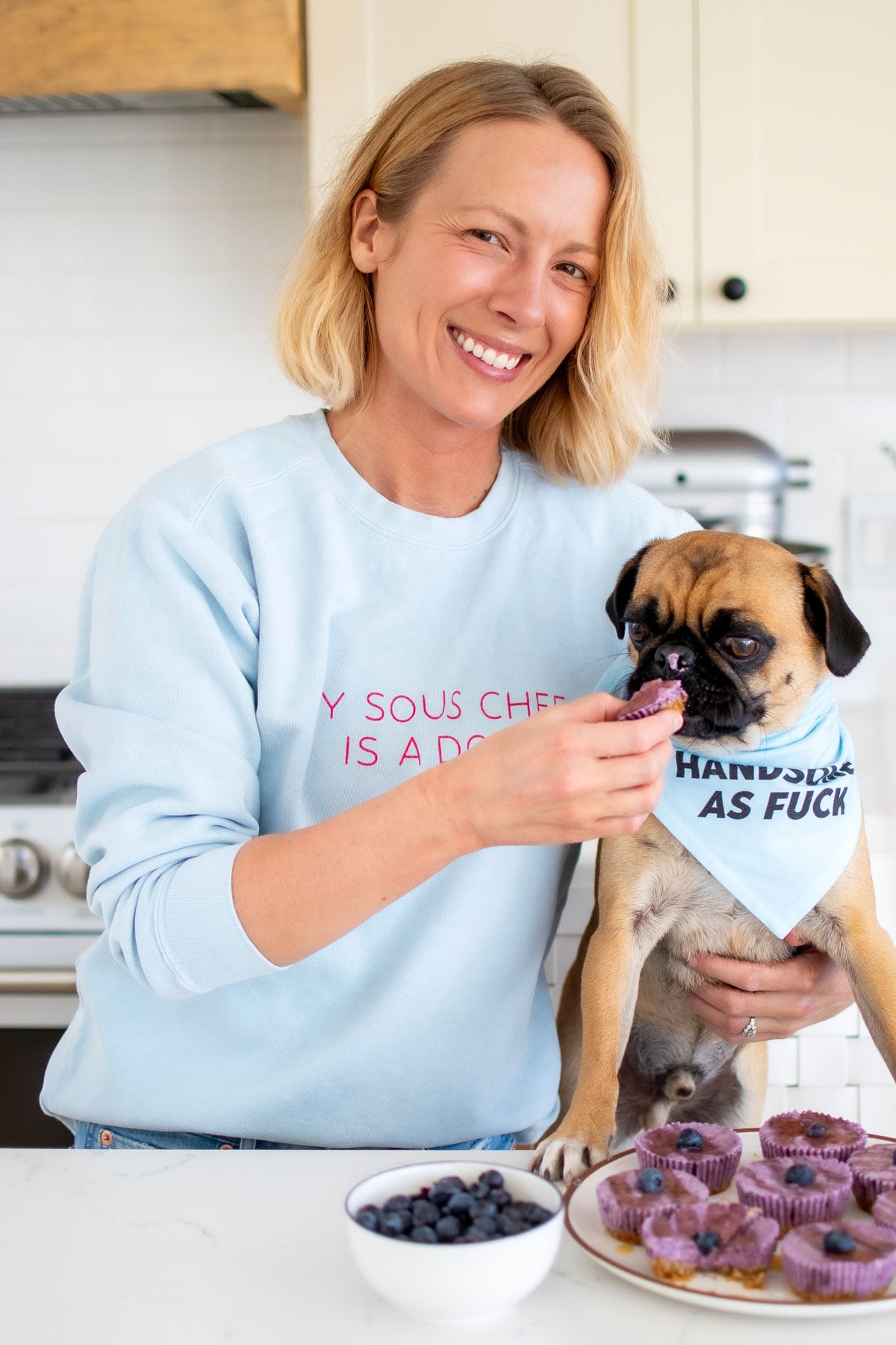 woman and pug in the kitchen, woman feeding pug blueberry cheesecake from a plate.