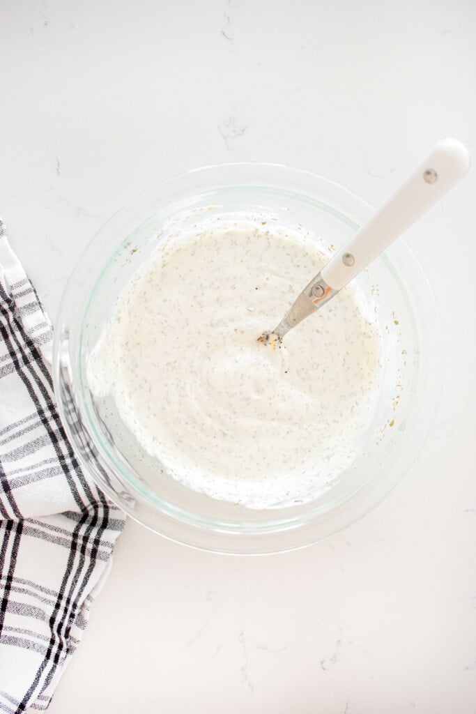 dill dip in a glass mixing bowl with a spoon on a marble counter.