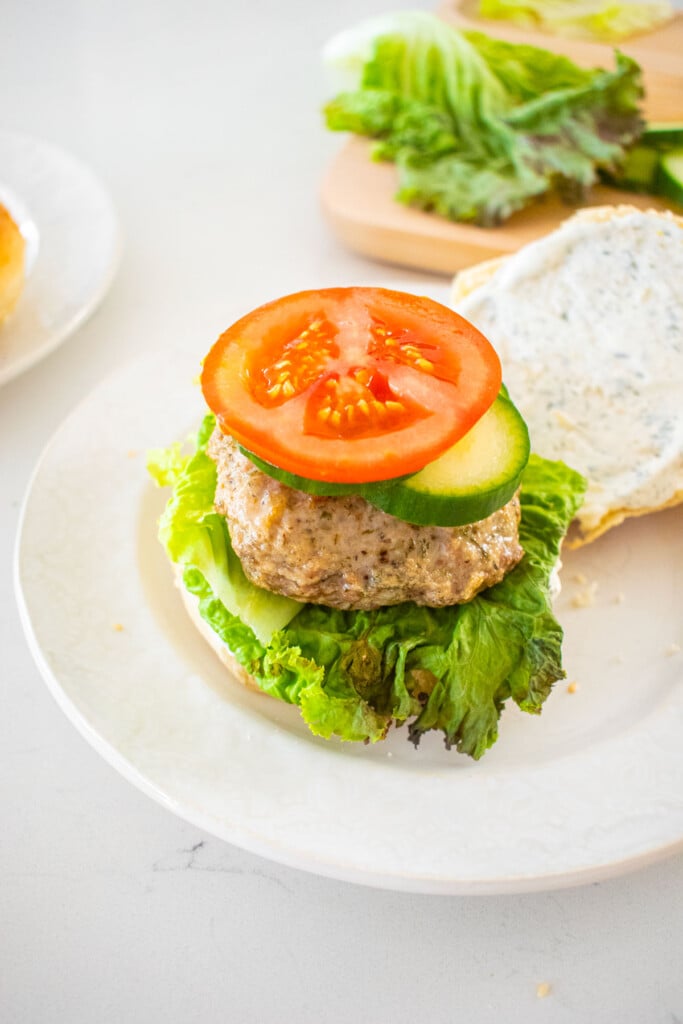 turkey and lamb burger being assembled on a bun with lettuce, tomato and cucumbers on a marble counter.