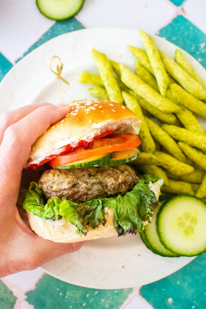 turkey lamb burger with dill sauce being grabbed with a hand on on plate on a green tile table.