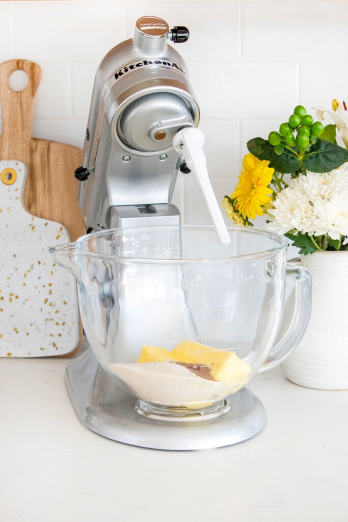 silver stand mixer with a glass mixing bowl and butter sugar and vanilla in it on a marble counter.
