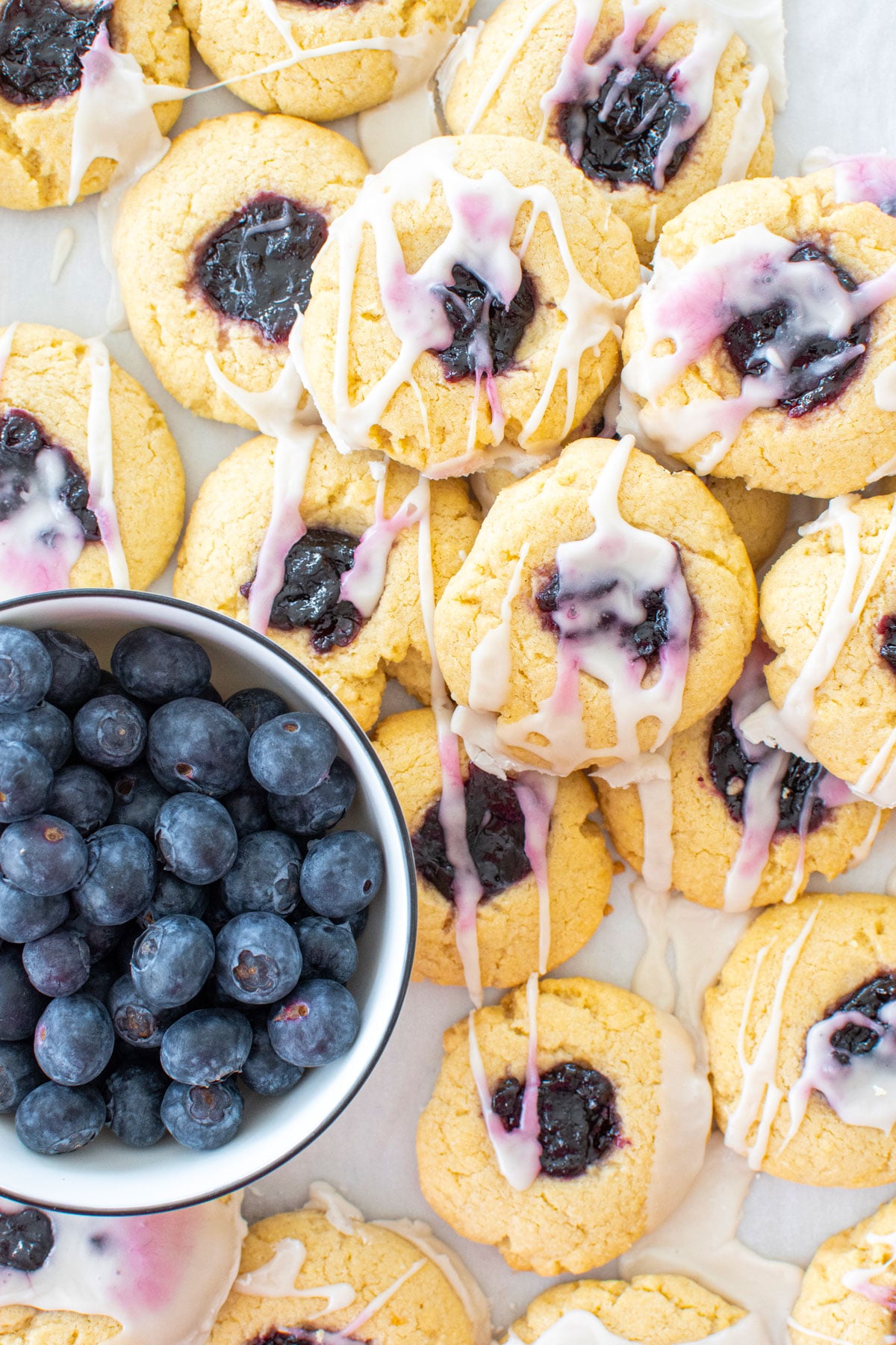 blueberry jam thumbprint cookies on a baking sheet with glaze and fresh blueberries.