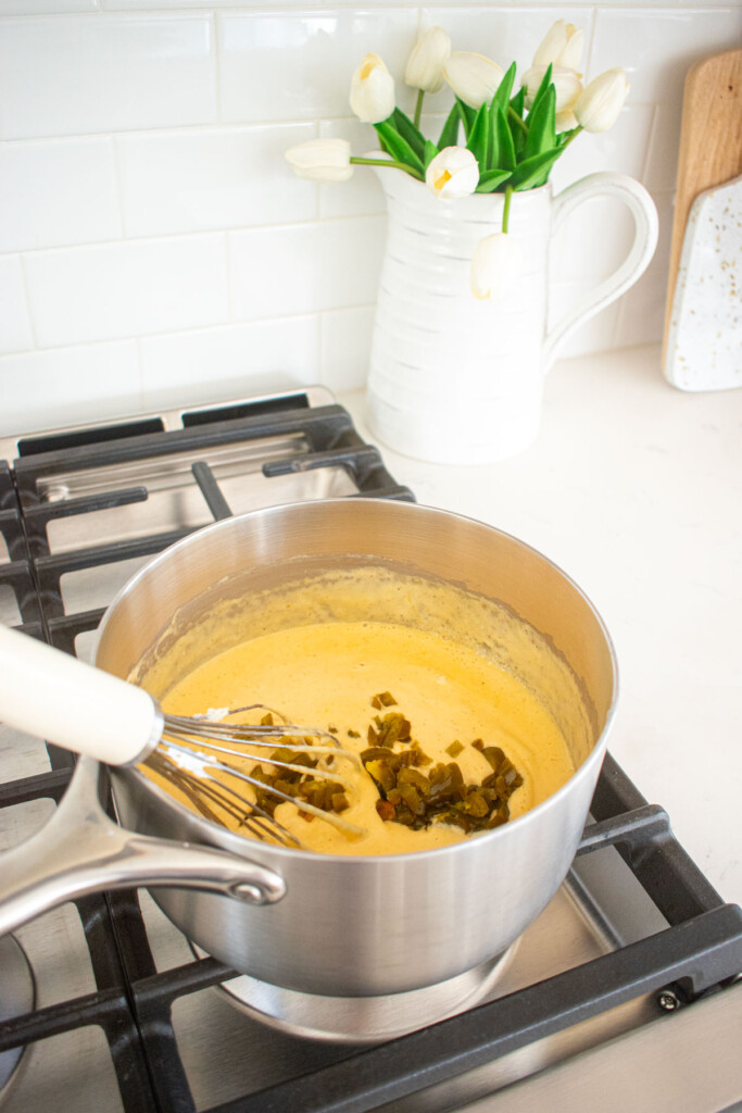 jalapenos being added to bone broth queso dip being stirred with a whisk in a pot on the stovetop.