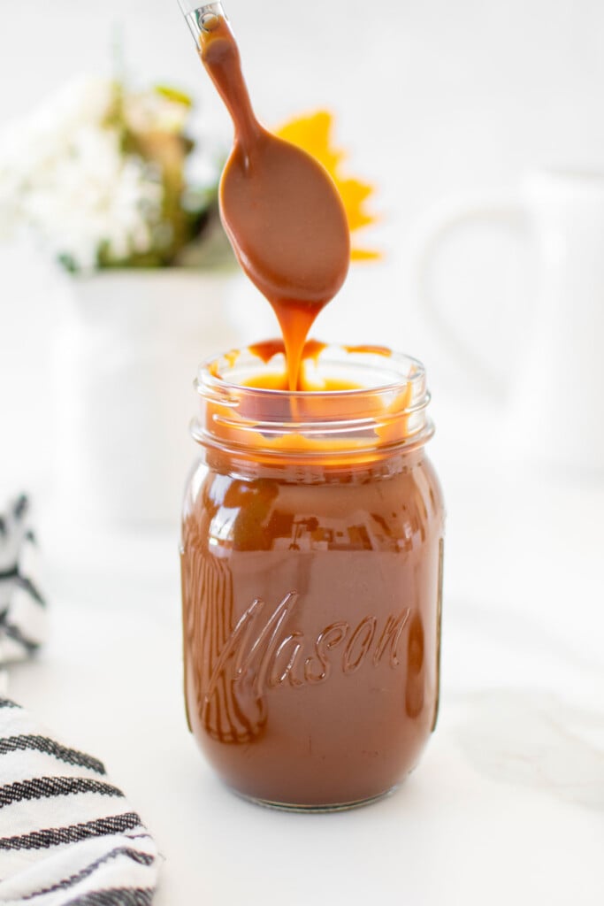 homemade true caramel sauce in a mason jar with a spoon dripping with caramel being taken out.
