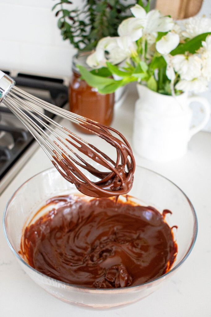 chocolate ganache on a whisk coming out of a glass mixing bowl on a white marble counter with fresh flowers.