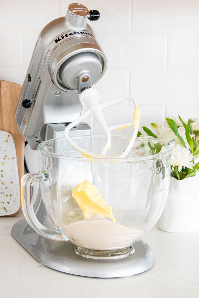 butter, sugar and vanilla extract in a glass mixing bowl on a stand mixer on a white marble counter with flowers.