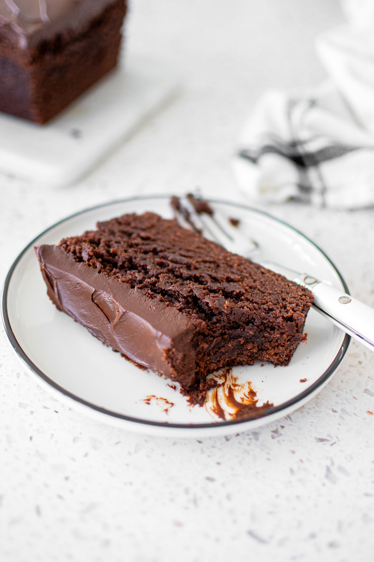 chocolate pound cake with chocolate ganache frosting on a plate with a bite taken out on a marble counter.