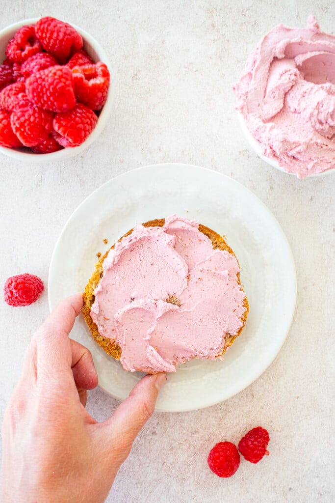 raspberry cream cheese spread on a bagel on a white plate, a woman grabbing it.