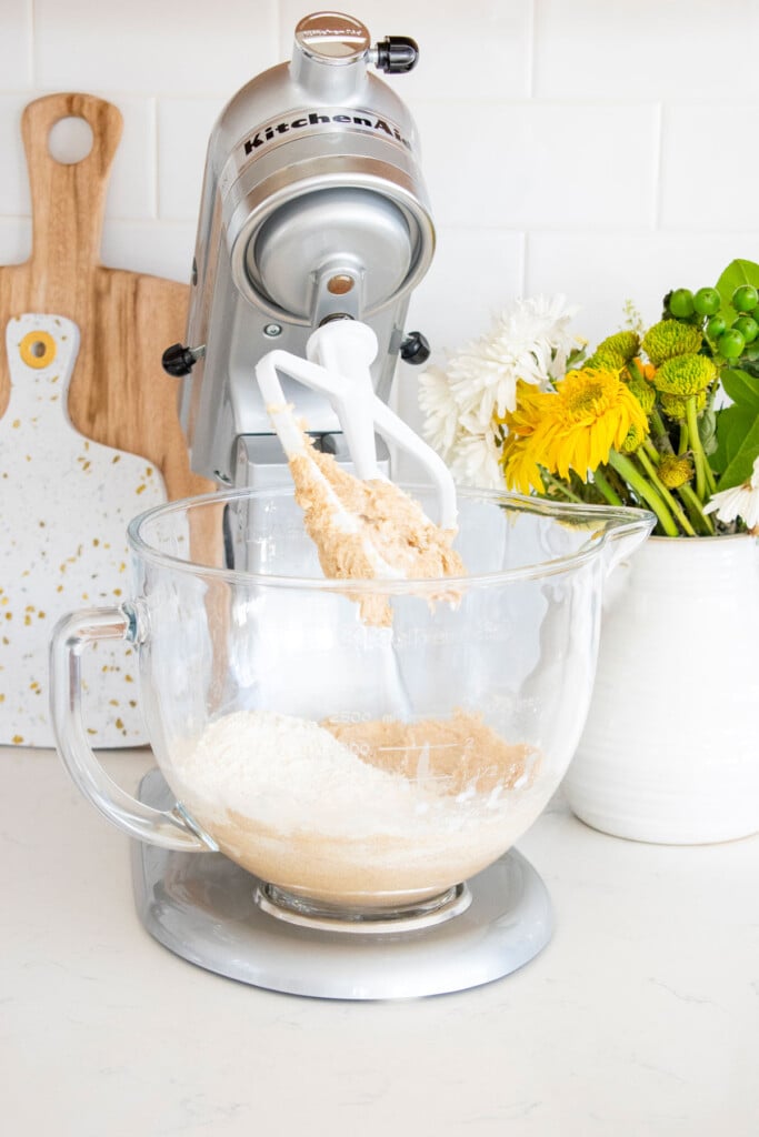 ingredients for vegan cookie dough in a stand mixer on a white marble counter with a vase of flowers.