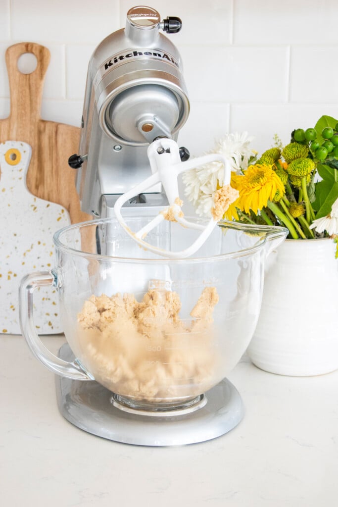 vegan cookie dough in a stand mixer on a white marble counter with a vase of flowers.