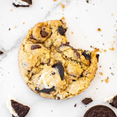 vegan oreo cookie with a bite taken out on a white marble counter with broken oreo pieces all around.