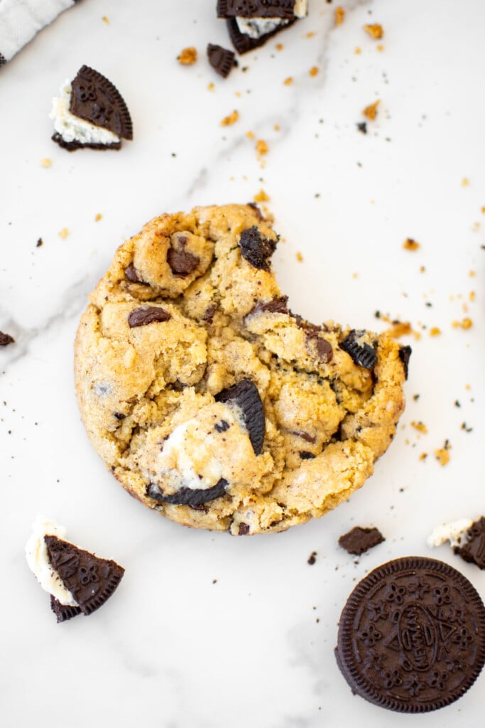 vegan oreo cookie with a bite taken out on a white marble counter with broken oreo pieces all around.
