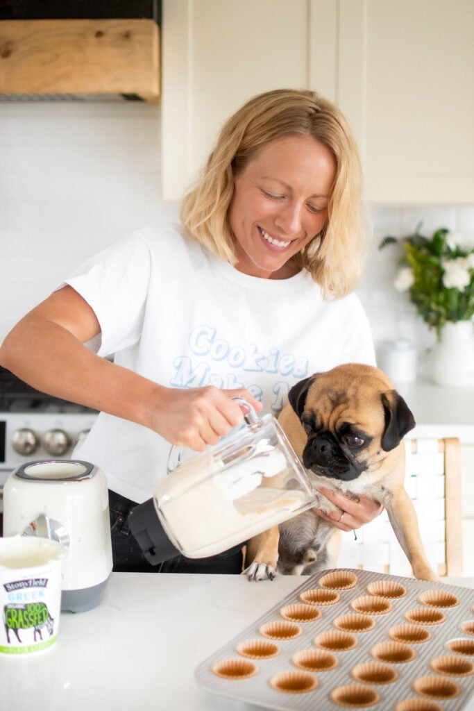 woman pouring frozen peanut butter dog treats batter into cupcake pan with her pug helping at kitchen counter.