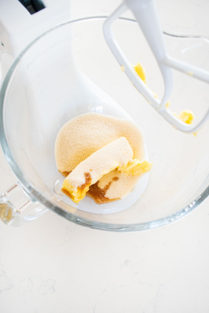 glass mixing bowl with butter sugar and vanilla extract on a white marble counter.