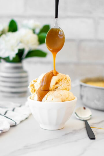 small bowl of caramel ice cream being drizzled with caramel sauce on a white marble counter with fresh flowers.