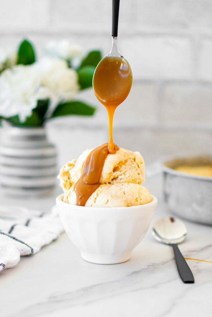 small bowl of caramel ice cream being drizzled with caramel sauce on a white marble counter with fresh flowers.