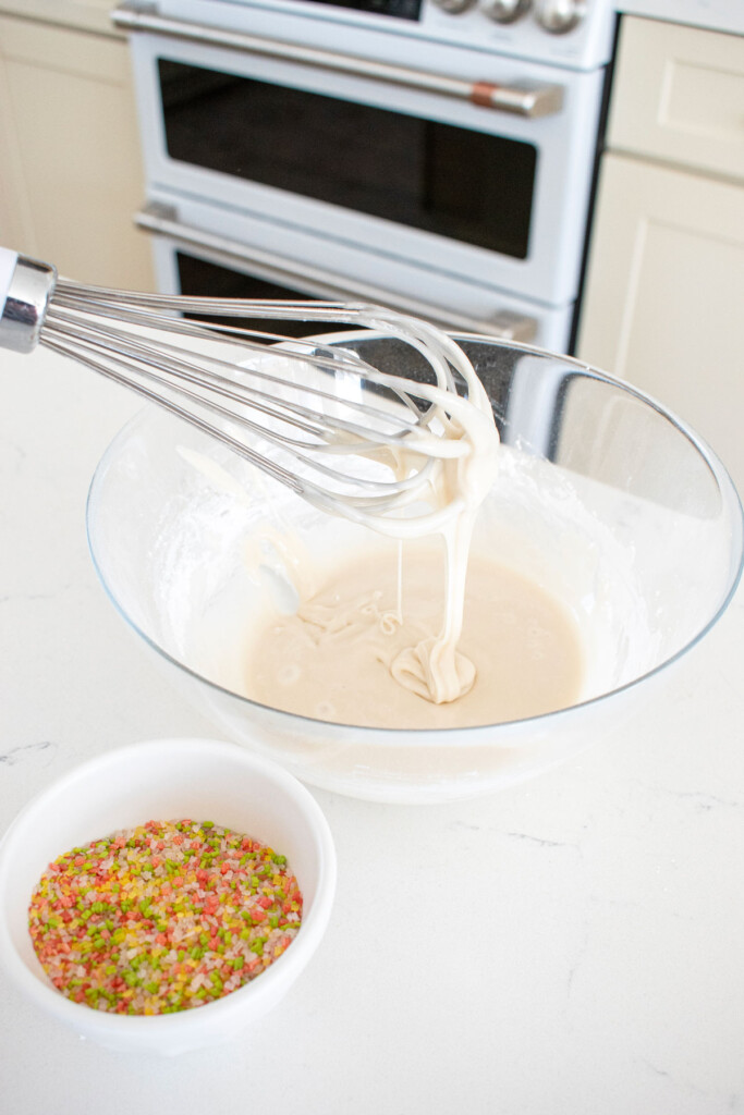 glaze being mixed in a glass bowl with a whisk on a white marble countertop.