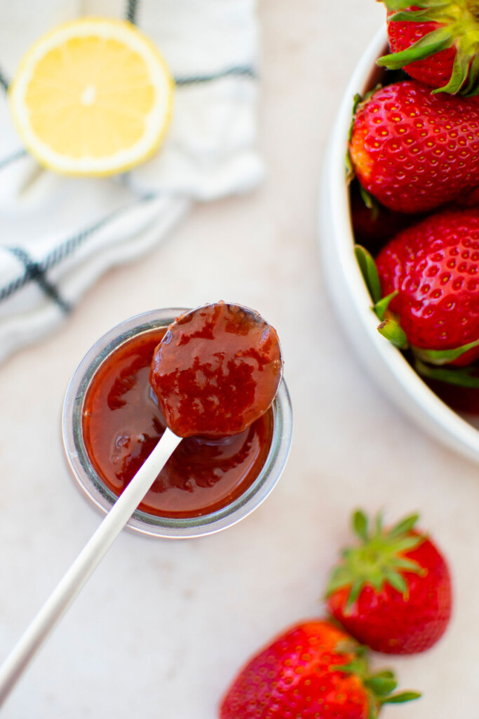 strawberry preserves on a spoon on top of a jar with a lemon and fresh strawberries.