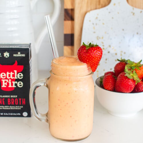 tropical bone broth protein smoothie with a fresh strawberry on the glass on a white marble counter with fresh strawberries.