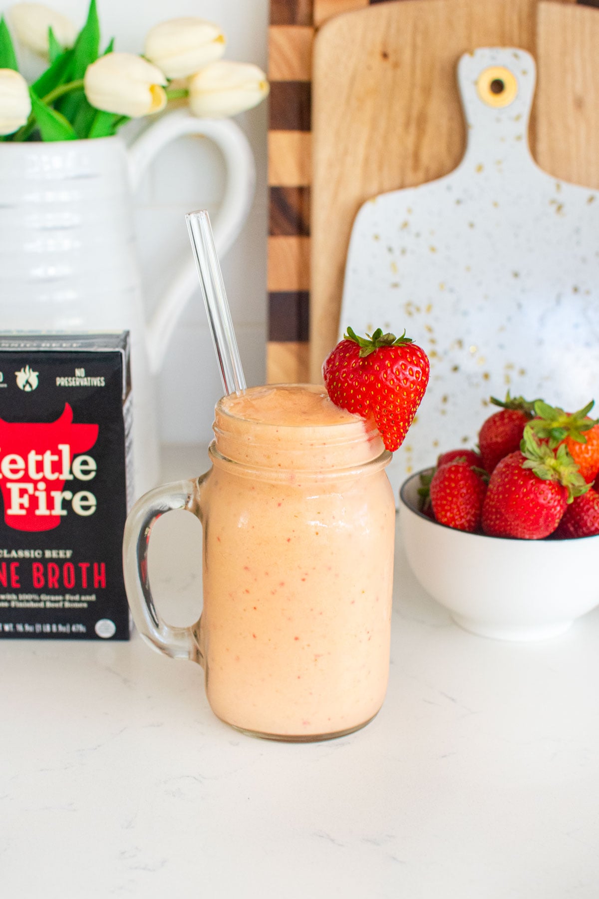 tropical bone broth protein smoothie with a fresh strawberry on the glass on a white marble counter with fresh strawberries.