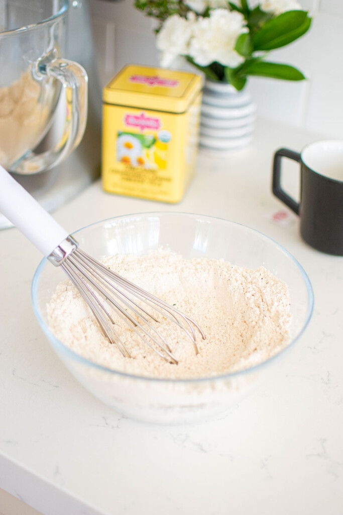 dry ingredients being mixed together in a glass mixing bowl with a whisk on a white marble counter.