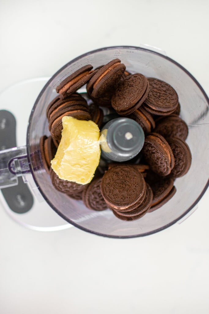 chocolate cookies and butter in a food processor on a white marble counter.