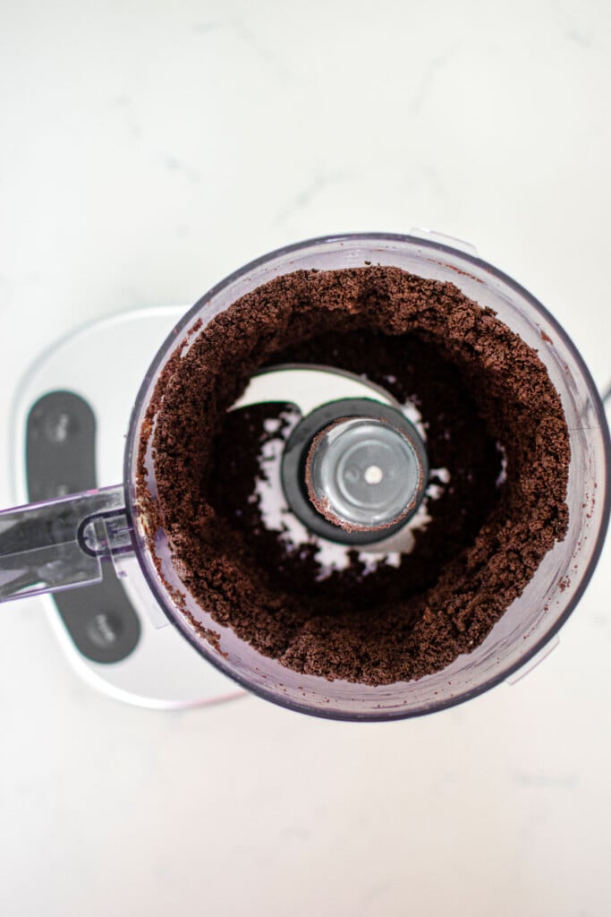 chocolate graham cracker crust in a food processor on a white marble counter.