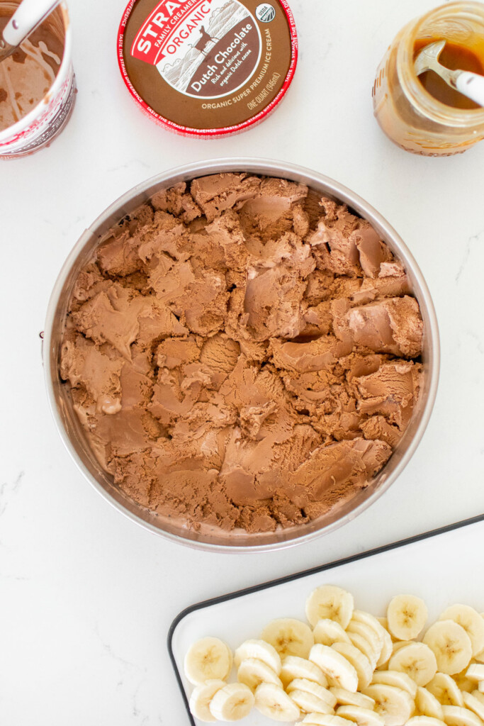 chocolate banana caramel ice cream pie being assembled on a white marble counter.
