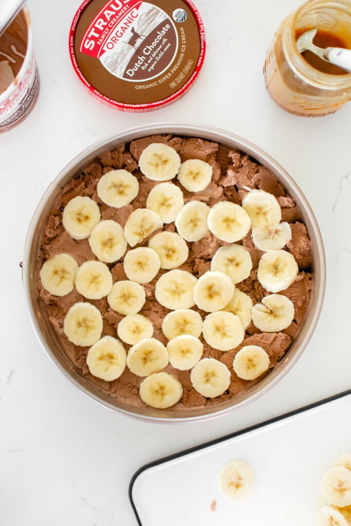 chocolate banana caramel ice cream pie being assembled on a white marble counter.