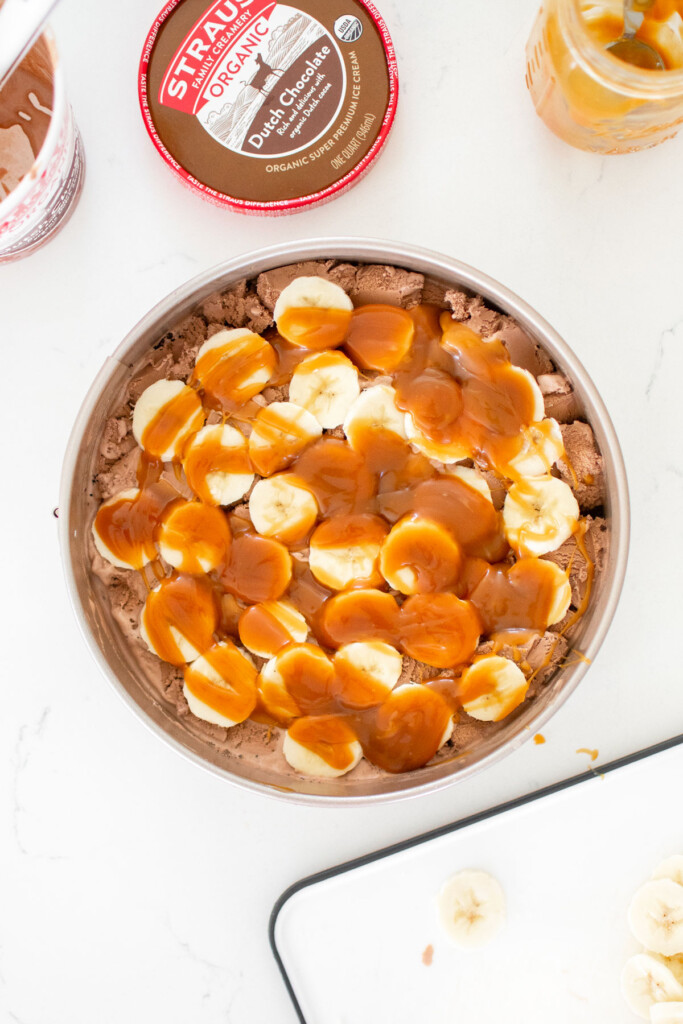 chocolate banana caramel ice cream pie being assembled on a white marble counter.