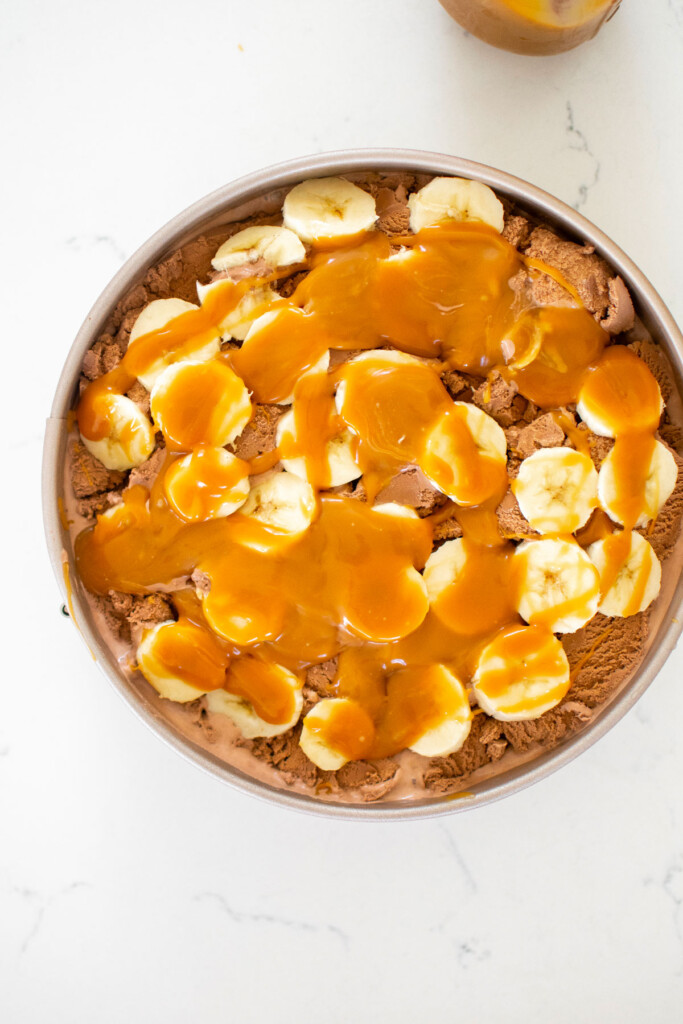 chocolate banana caramel ice cream pie being assembled on a white marble counter.