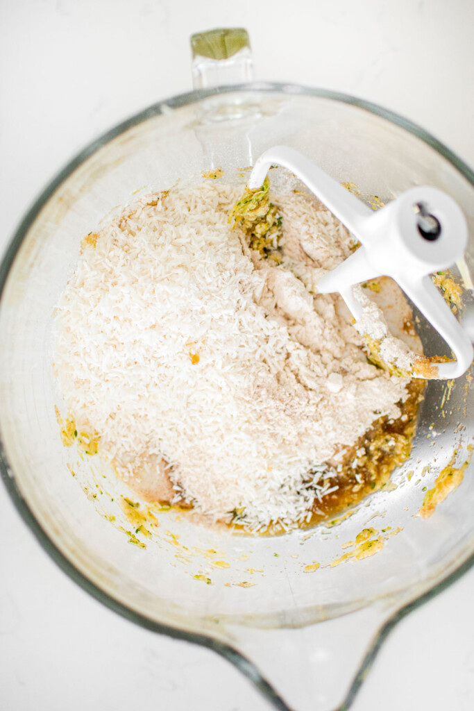 ingredients for eggless strawberry zucchini cake in a glass stand mixing bowl on a white marble counter.