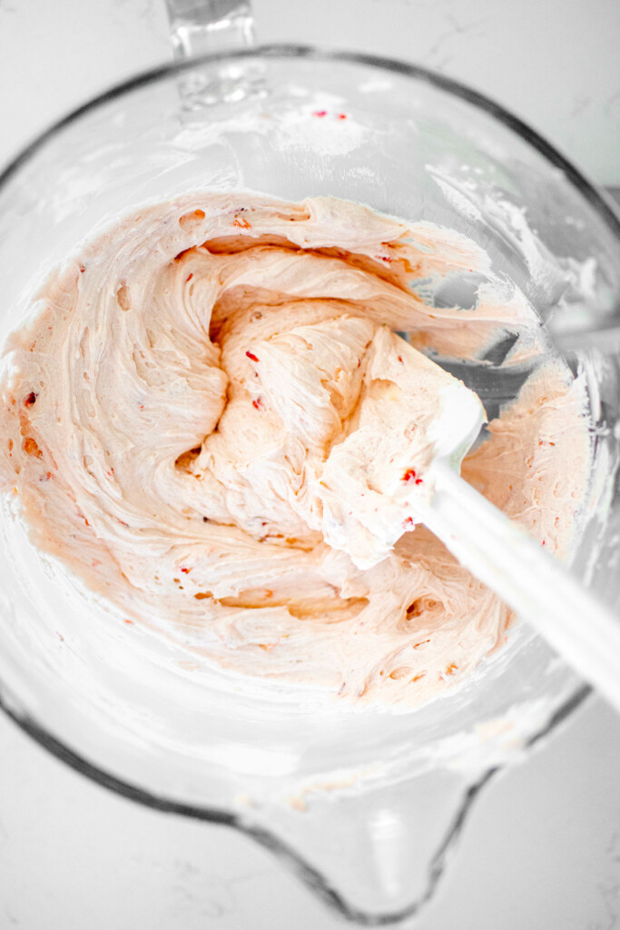 light pink strawberry buttercream in a glass mixing bowl with a spatula. ona white marble counter.