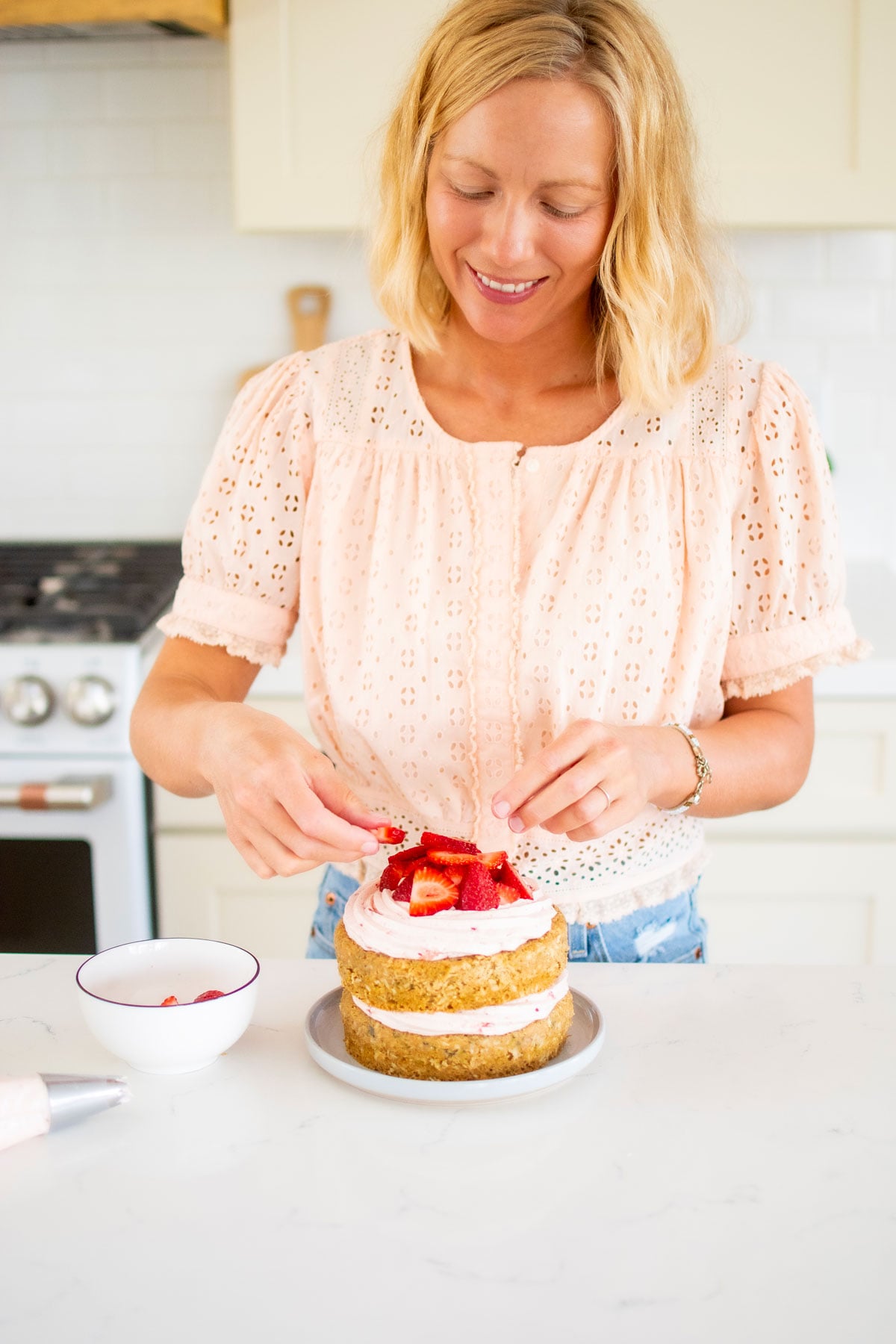 woman decorating eggless strawberry zucchini cake with fresh strawberries on a cake plate in the kitchen.