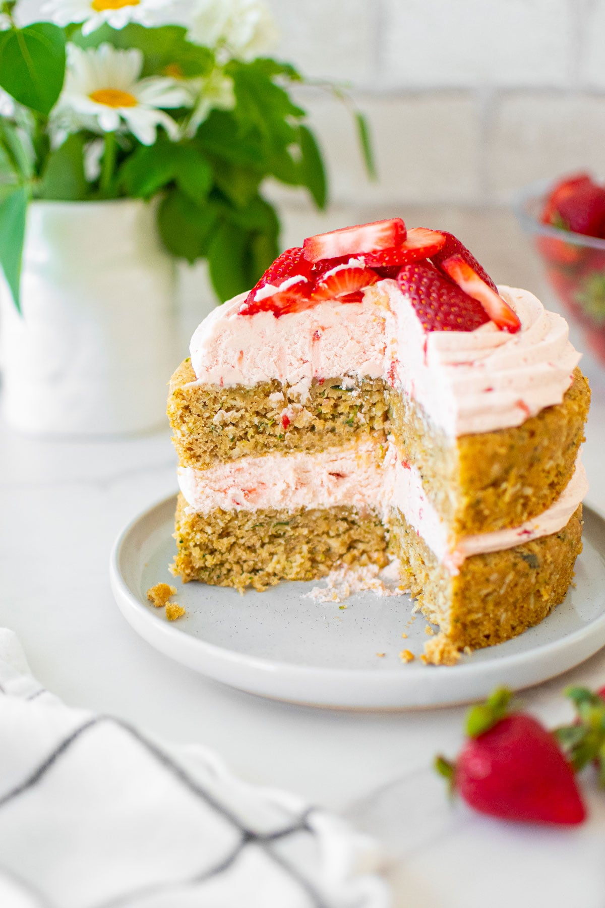 eggless strawberry zucchini cake with fresh strawberries on top cut into on a cake plate on a marble table with fresh flowers.