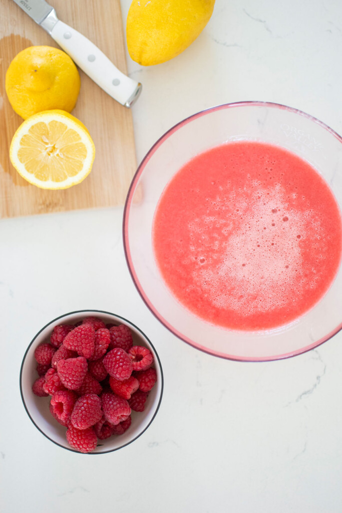 strained lemon and raspberry juice in a glass bowl with lemons and raspberries on a white marble countertop.
