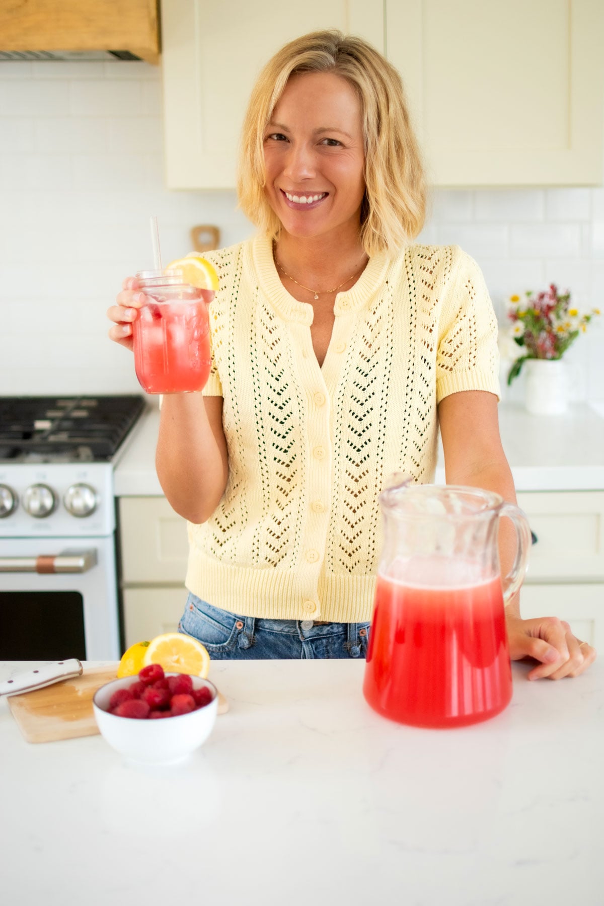 woman holding a glass of organic pink lemonade next to the pitcher at the kitchen counter.