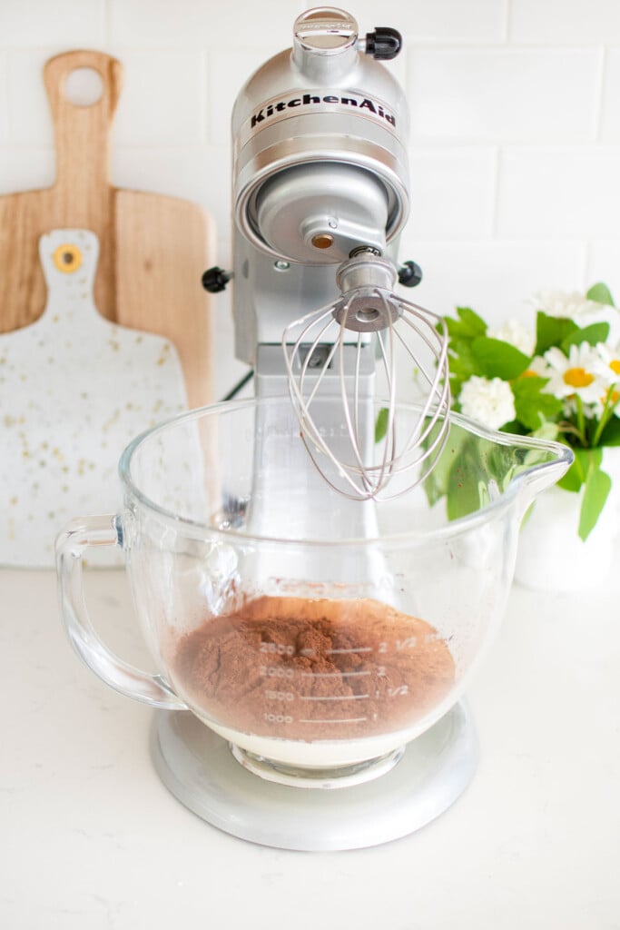 cocoa and whipping cream in glass stand mixer bowl on a white countertop.