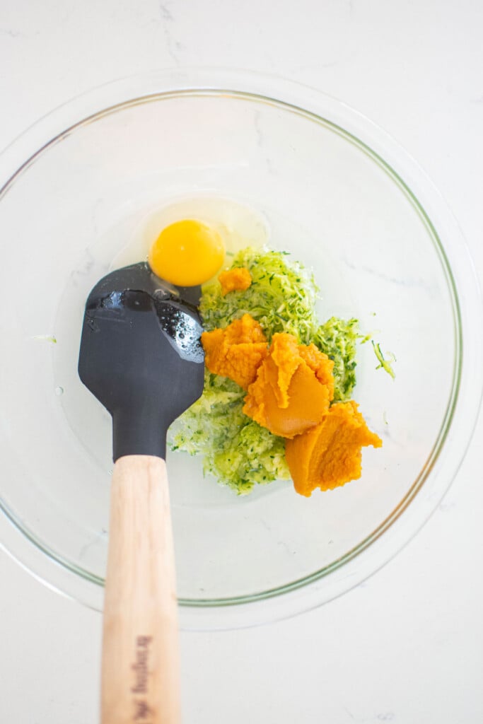 grated zucchini, eggs, and pumpkin in a glass mixing bowl on a white countertop.