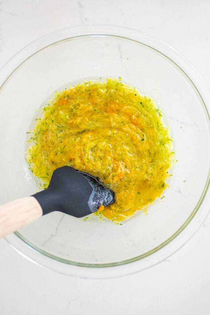 grated zucchini, eggs, and pumpkin mixed together with a spatula in a glass mixing bowl on a white countertop.