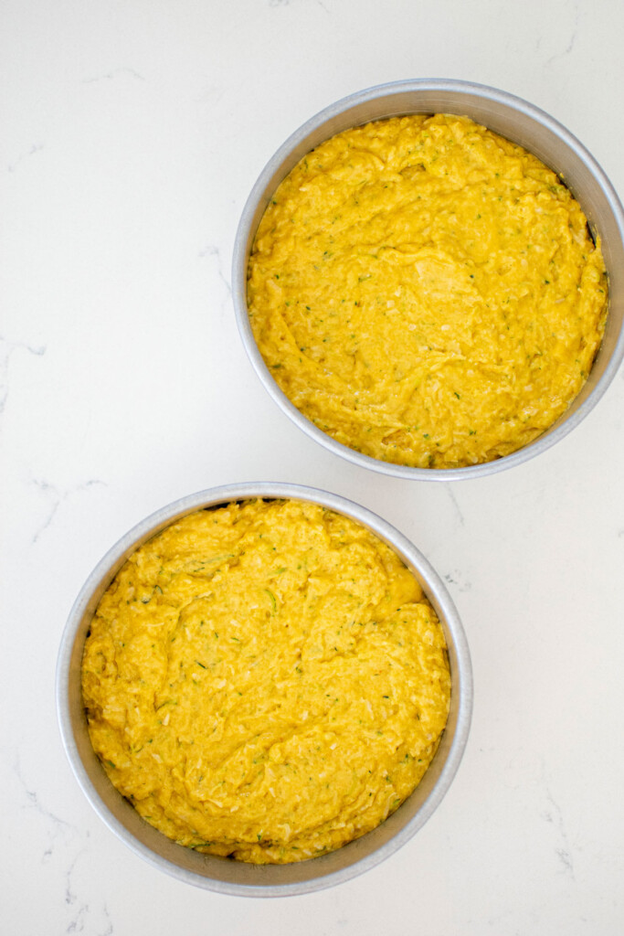 zucchini dog cake batter in two cake pans on a white marble countertop.