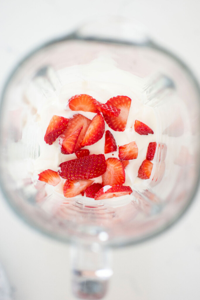 strawberries and greek yogurt in a glass blender on a white marble countertop.