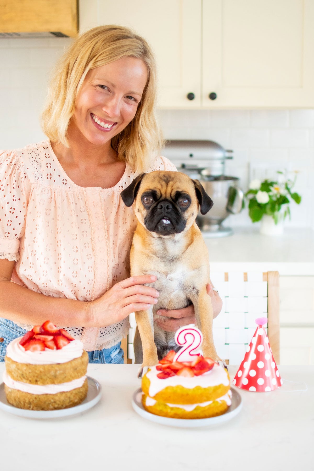 woman and pug at kitchen counter with two strawberry zucchini birthday cakes and a birthday hat.