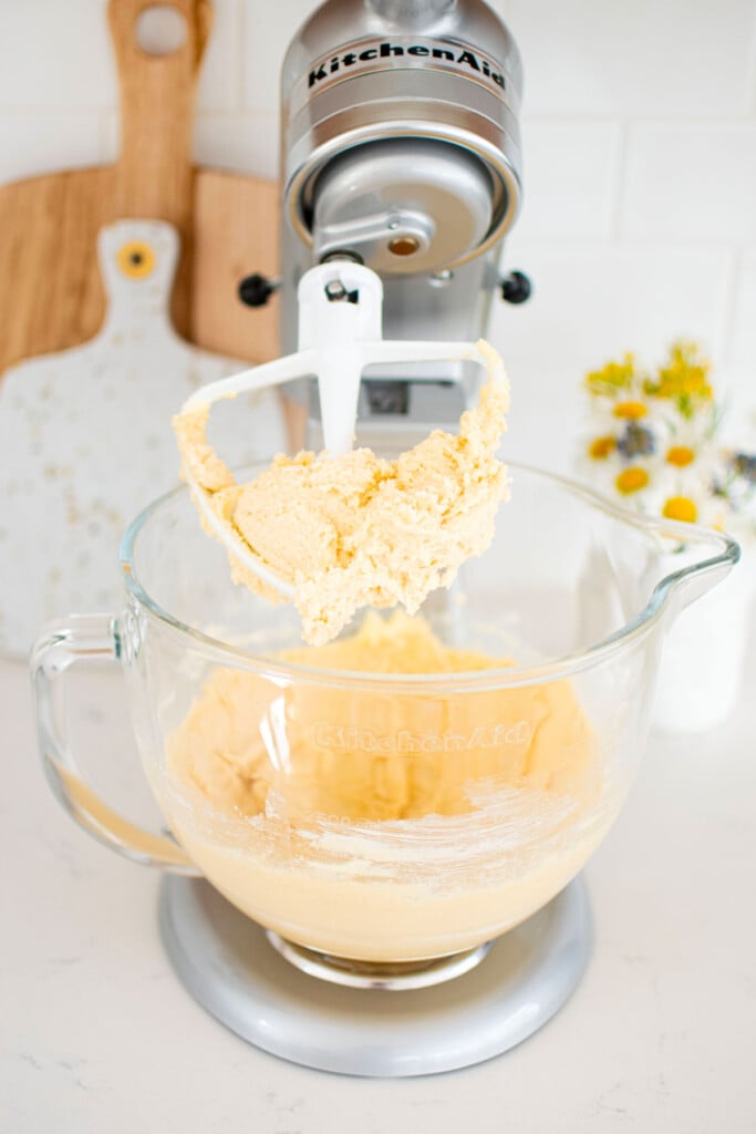 butter, sugar and vanilla creamed together in a silver stand mixer on a white marble counter with a vase of flowers.
