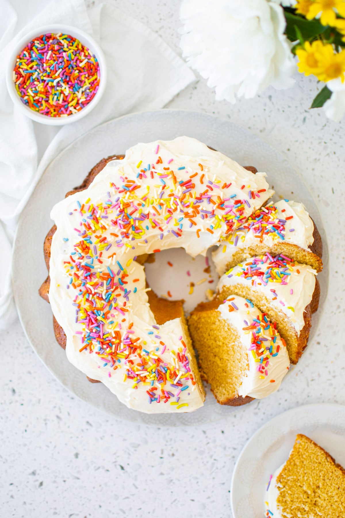 birthday bundt cake with rainbow sprinkles on a cake plate cut into slices with fresh flowers.