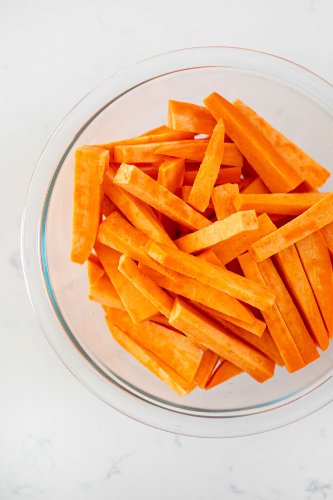 peeled and cut sweet potato fries in a glass mixing bowl on a white marble countertop.
