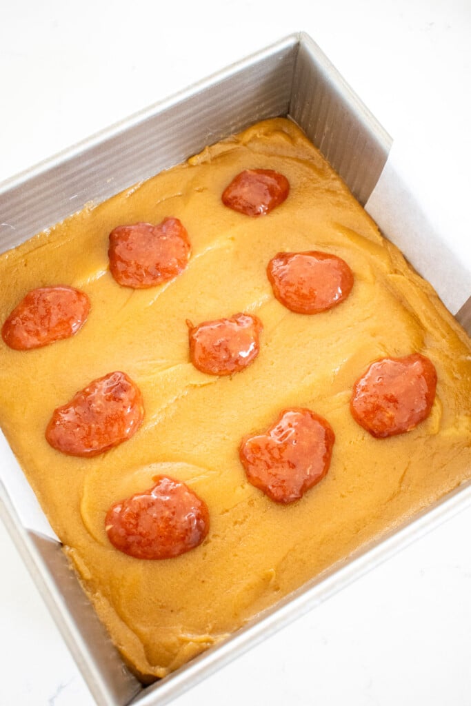 strawberry jam in dollops on top of olive oil brownie batter in a square pan on a white marble counter.