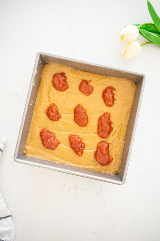 olive oil blondie batter in a square baking pan with dollops of strawberry jam on a marble counter.