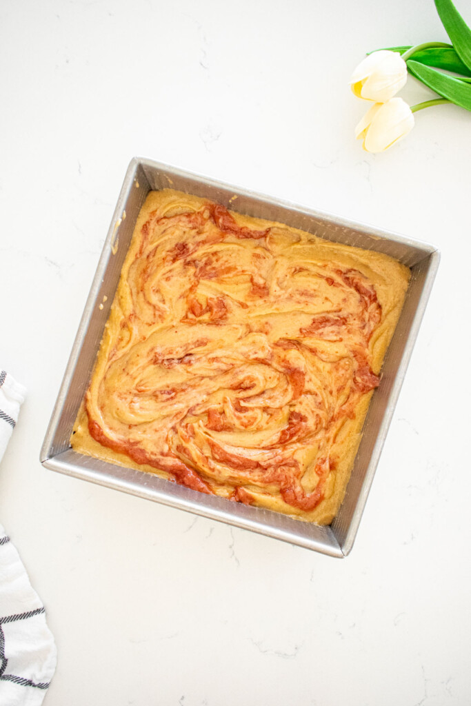 olive oil blondie batter with strawberry jam swirl in a square baking pan on a marble counter.